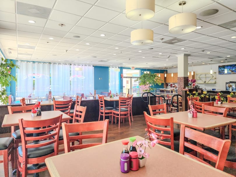 Spacious, brightly lit restaurant dining room with many empty tables and wooden chairs, and blue accent walls.