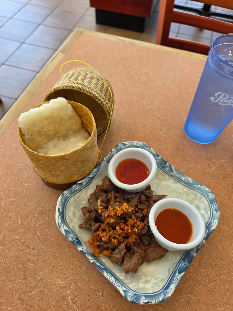 Fried beef with crispy garlic, two dipping sauces, sticky rice in a bamboo basket, and a drink.