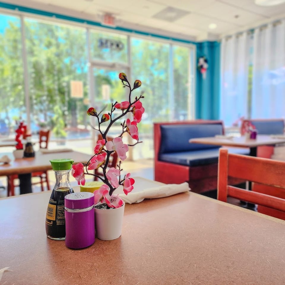 A restaurant interior with a table in the foreground, featuring a soy sauce bottle and a pink floral decoration.