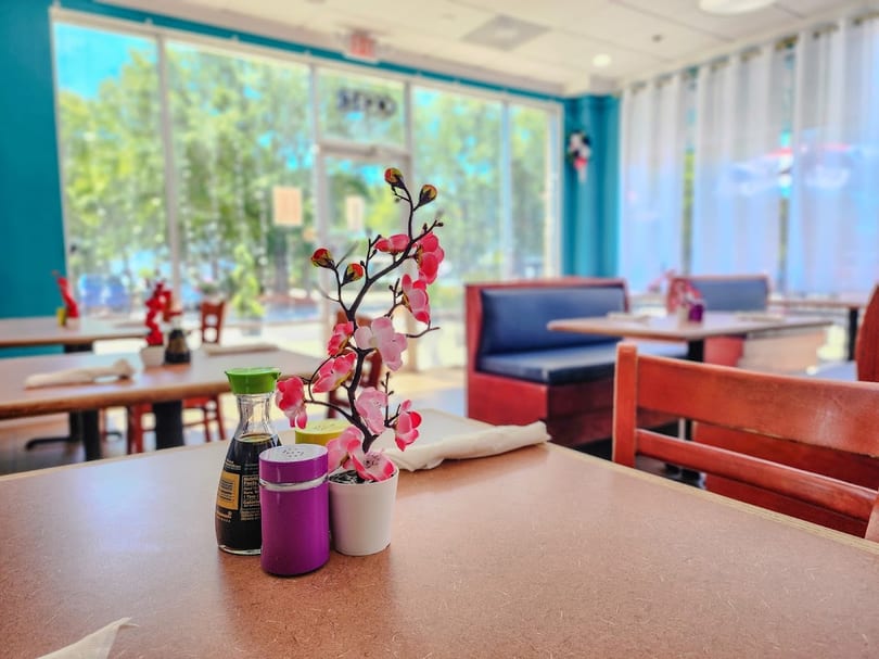 A restaurant interior with a table in the foreground, featuring a soy sauce bottle and a pink floral decoration.