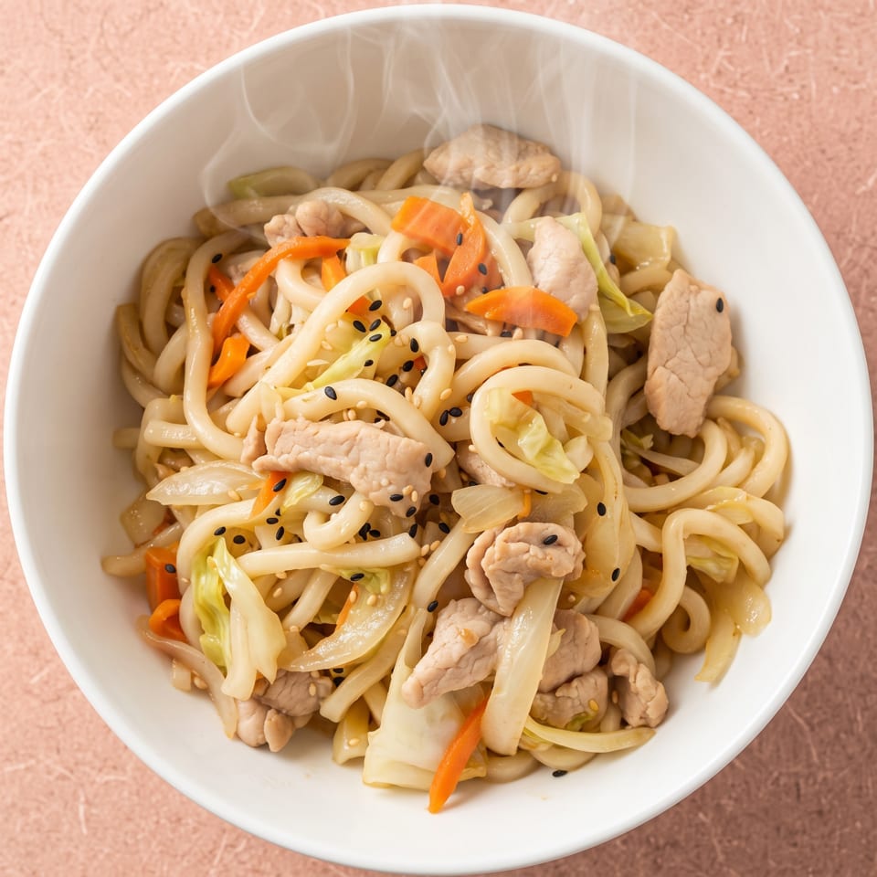 Steaming udon noodles with pork, carrots, cabbage, and sesame seeds in a white bowl.